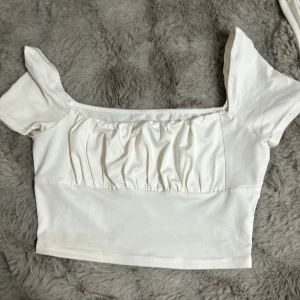 Vit kortärmad topp - Säljer en söt vit topp med korta puffärmar och rynkad detalj framtill. Toppen är croppad och har en mjuk och bekväm passform. Perfekt för varma dagar!