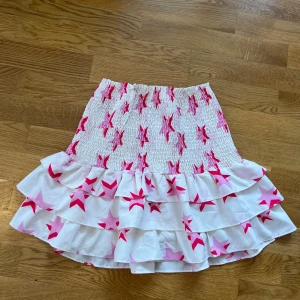 Vit och rosa kjol med stjärnmönster - Loavies vit kjol med rosa stjärnor och volanger. Den har en elastisk midja för bekväm passform. Finns lite fläckar men annars fin