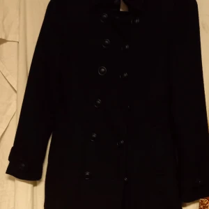 Svart fodrad finkappa från Bik Bok - Snygg svart fodrad finkappa från Bik Bok i storlek XS. Den har en klassisk dubbelknäppt design med långa ärmar och är tillverkad i en blandning av polyester, viskos och ull. Perfekt för kyligare dagar.