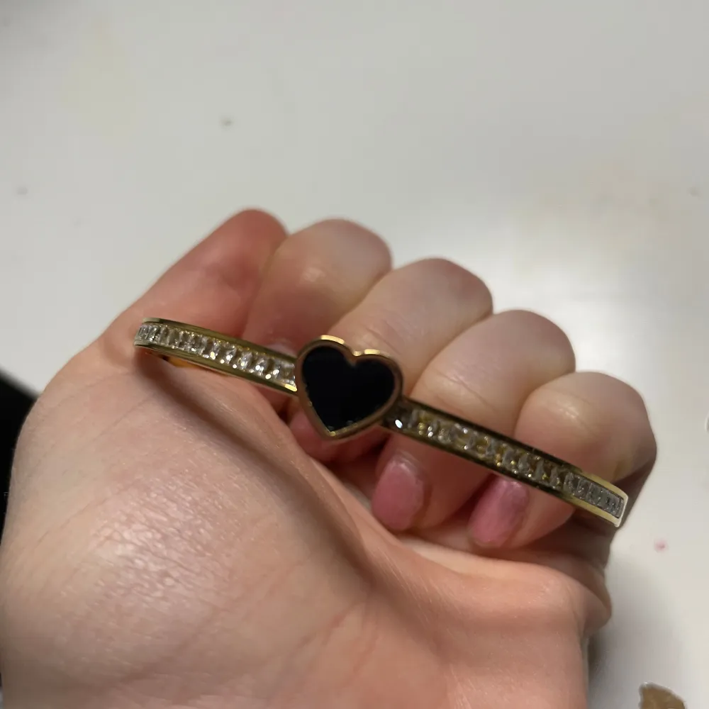 Elegant guldarmband med ett svart hjärta i mitten. Armbandet är dekorerat med glittrande stenar längs hela bandet, vilket ger en lyxig känsla. Perfekt för att lägga till en touch av glamour till din outfit.. Asusteet.