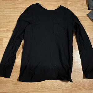 Svart långärmad tröja - Säljer en stilren svart långärmad tröja med en liten bröstficka. Perfekt för en enkel och avslappnad look. Tröjan har en normal passform och är tillverkad i ett mjukt material. 