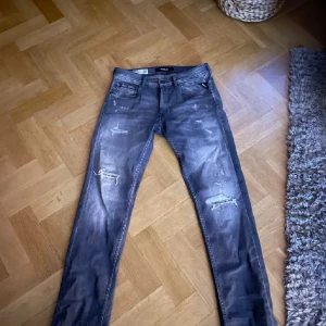 Grå jeans från Replay - Snygga grå jeans från Replay med slitna detaljer och en klassisk femficksdesign. De har en straight passform och är perfekta för en avslappnad stil. Märkeslogga på baksidan av midjan.