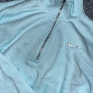 Ljusblå tröja med dragkedja från Nike - Säljer en ljusblå tröja från Nike med halv dragkedja och hög krage. Perfekt för en sportig look eller avslappnad stil. Den har en enkel design med det klassiska Nike-loggan på bröstet.