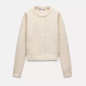Beige stickad tröja från Zara - Säljer en stilren beige stickad tröja från Zara. Tröjan har en enkel design med en subtil vit linje framtill och ribbade kanter vid ärmslut och nederkant. Perfekt för en avslappnad look.