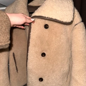 Beige teddyjacka med knappar - Säljer en mysig beige teddyjacka med stora knappar framtill. Jackan har en klassisk krage och är perfekt för kyligare dagar. Den är långärmad och har en mjuk, fluffig yta som ger en trendig look. Från arket, nypris 2700kr. Inga tecken på användning eller defekter 