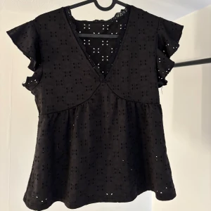 Svart blus med volangärmar - Säljer en elegant svart blus med v-ringning och volangärmar. Blusen har ett fint broderat mönster och är perfekt för både vardag och fest. Den är tillverkad i ett lätt och luftigt material. Använd ca 5 gånger, nästan som oanvänd, mycket bra skick😊