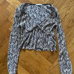 Blommig omlottopp - Fin blommig omlottopp. Toppen har långa ärmar och ett fint blommönster i lila, blått och svart. Har ett svalt skönt tyg. Perfekt för vår och sommar!