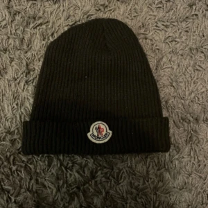  mössa från Moncler - Snygg svart stickad mössa från Moncler med uppvikt kant och det klassiska Moncler-emblemet framtill. Perfekt för kyliga dagar.