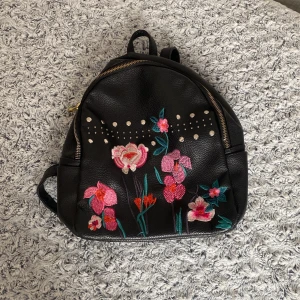 Svart ryggsäck med blommigt broderi från OVS - Snygg svart ryggsäck i syntetmaterial med färgglatt blommigt broderi och dekorativa nitar. Perfekt för att ge din outfit en unik touch. Ryggsäcken har dragkedja och justerbara axelremmar för bekväm passform.