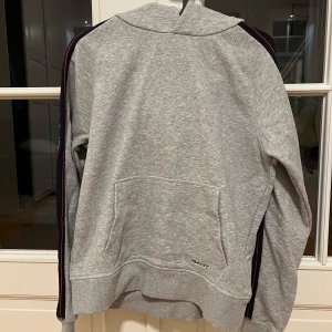 Grå hoodie från GANT - Snygg grå hoodie från GANT med logotyp längs ärmarna. Perfekt för en avslappnad stil. Den har en klassisk design med ribbade muddar och en bekväm passform.