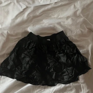 Svart kortkjol - Snygg svart kortkjol med en glansig finish. Perfekt för att ge en stilren look. Kjolen har en elastisk midja för bekväm passform.