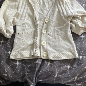 Vit kofta från H&M - Säljer en söt vit kofta från H&M med stora knappar framtill. Koftan har trekvartsärmar och ett fint ribbat mönster vid ärmslut och nederkant. Perfekt för en stilren look!