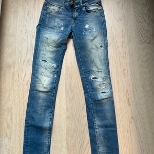 Blå jeans från Replay - Snygga blå jeans från Replay med slitna detaljer och en klassisk femficksdesign. De har en knappgylf och är perfekta för en avslappnad stil. Märkesdetaljer syns på fickan och baksidan.