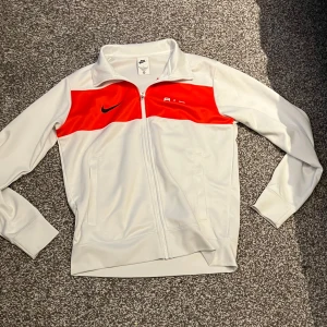Vit kofta från nike  - Säljer en snygg vit och röd kofta från Nike med dragkedja och långärmade ärmar. Koftan har en röd panel över bröstet med Nike-loggan och texten 'AIR'. Perfekt för en sportig look!