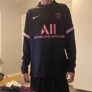 Svart och rosa fotbollströja från Nike - Snygg svart fotbollströja från Nike med rosa detaljer och Paris Saint-Germain-logga. Tröjan har långa ärmar och en halv dragkedja framtill. Perfekt för fotbollsfans som vill ha stil både på och utanför planen.