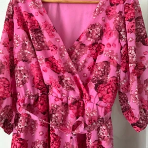Rosa blommig klänning från Happy Holly - Säljer en superfin rosa klänning från Happy Holly med blommigt mönster i olika nyanser av rosa och rött. Klänningen har en omlott-design med v-ringning och knytband i midjan. Perfekt för en färgglad look!