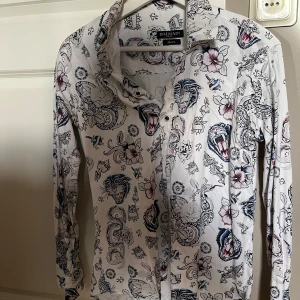 Vit mönstrad skjorta från Balmain - Snygg vit skjorta från Balmain med ett unikt mönster av djur och blommor i svart och rött. Skjortan har långa ärmar och knappar framtill. Perfekt för dig som vill sticka ut med en cool och stilren look.