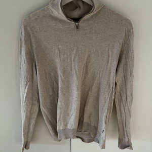 Beige tröja med dragkedja halfzip  - Säljer en stilren beige tröja med dragkedja vid halsen. Tröjan har långa ärmar och ribbade muddar vid ärmslut och nederkant. Perfekt för en avslappnad look.