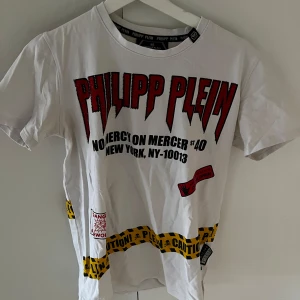 Vit t-shirt från Philipp Plein - Cool vit t-shirt från Philipp Plein med stora röda bokstäver och detaljer i svart och gult. Texten 'Mercer St' och en dödskalle finns på baksidan. Perfekt för en edgy look!