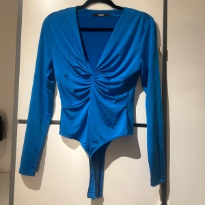 Blå långärmad body  - Snygg blå långärmad body med v-ringning och knytdetalj framtill. Materialet är mjukt och stretchigt för en bekväm passform.