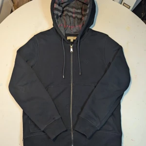 Svart hoodie från Burberry - Snygg svart hoodie från Burberry med dragkedja och klassiskt rutigt mönster i huvan. Tröjan har långa ärmar och en diskret broderad logga på bröstet. Perfekt för en stilren look.