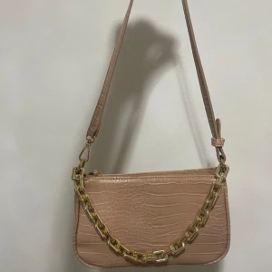 Beige axelväska med kedjedetalj - Snygg beige axelväska med krokodilpräglat mönster och en iögonfallande guldfärgad kedja. Väskan har en justerbar axelrem och stängs med dragkedja. Perfekt för att ge din outfit en trendig touch.
