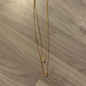 Guldigt dubbelt halsband med hjärta och sten - Elegant dubbelt halsband i guld med två kedjor. Det ena hänget är en klar sten och det andra är ett hjärta med en sten i mitten😁