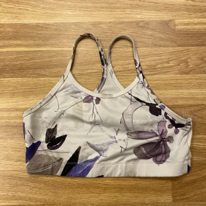 Blommig sporttopp från SOC - barn  - Säljer en blommig sporttopp från SOC i ljusa nyanser med lila och svarta blommor. Toppen har smala axelband och är tillverkad i ett mjukt och bekvämt material som håller dig torr under träning.