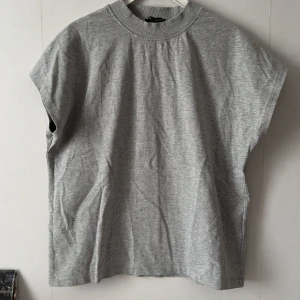 Grå topp från Zara - Säljer en stilren grå topp från Zara. Den har en ribbad krage och är ärmlös vilket gör den perfekt för en avslappnad look. Materialet är mjukt och bekvämt, den är i storlek för att den ska sitta lös på, använt endast en gång.