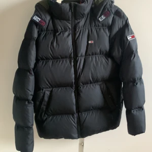 Svart pufferjacka från Tommy Jeans - Säljer en svart pufferjacka från Tommy Jeans med broderade logotyper på ärmarna och bröstet. Jackan har en hög krage och dragkedja framtill. Perfekt för kyliga dagar.