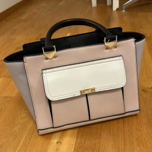 Elegant handväska i rosa och grått - Snygg handväska i en kombination av rosa och grått med svarta handtag. Väskan har en vit ficka framtill med guldfärgade detaljer. Perfekt för att ge din outfit en stilfull touch.