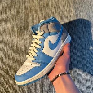 Air Jordan 1 Mid - Säljer ett par riktigt schyssta Air Jordan 1 Mid i storlek 41. Skorna är knappt använda och alltid burits med crease-skydd. De är även impregnerade för att stå emot smuts och fukt. Skicket är mycket bra, inga större tecken på slitage. Originalskosnören och sula i fint skick.  Perfekta för dig som vill ha ett par stilrena och fräscha sneakers i en snygg blå/vit färgkombination nu till våren! Ordinarie pris: 3200kr Det är bara att höra av sig vid frågor!