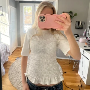 Vit smockad topp - Säljer en vit smockad topp med korta ärmar och en volangdetalj vid nederkanten. Toppen har en tight passform och är perfekt för en stilren look.