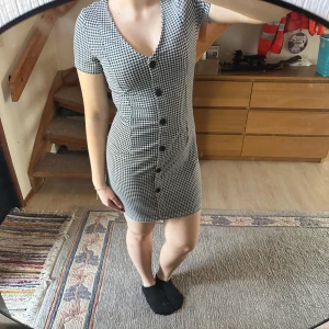 Svartvit rutig klänning - Säljer en snygg svartvit rutig klänning med korta ärmar och knappar framtill. Klänningen har en v-ringad design och en tight passform som framhäver figuren. Perfekt för en stilren look!
