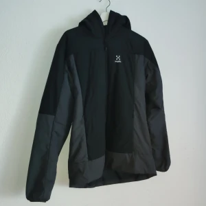 Svart vindjacka från Haglöfs - Säljer en svart vindjacka från Haglöfs med huva och dragkedja. Jackan har en stilren design med två sidofickor och är perfekt för utomhusaktiviteter. Den är tillverkad i ett slitstarkt material som skyddar mot vind.