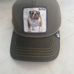 Olivgrön keps med bulldogmotiv -  Den är använd ett fåtal gånger Snygg olivgrön keps med broderad bulldog på framsidan och texten 'BUTCH'. Kepsen har ett nätmaterial baktill för ventilation och justerbar passform med ett grönt spänne. Perfekt för en avslappnad stil.