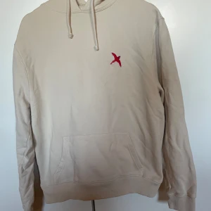 Beige hoodie från Axel Arigato - Säljer en stilren beige hoodie från Axel Arigato med en röd broderad fågel på bröstet. Näst intill aldrig använd! Storlek S