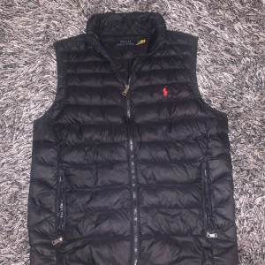 Polo Ralphlauren väst - Snygg svart dunväst från Polo Ralph Lauren med dragkedja och röd logotyp på bröstet. Västen är packbar och perfekt för kyliga dagar. Den har en stilren design med quiltade detaljer. Skick 9/10. Inga defekter