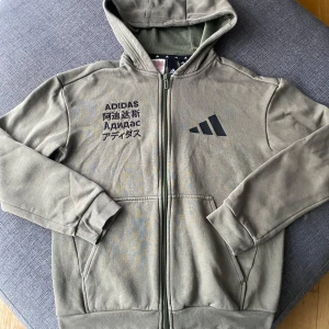 Olivgrön hoodie från Adidas - Säljer en olivgrön hoodie från Adidas med dragkedja och stor logga på ryggen. Tröjan har långa ärmar och en bekväm huva. Perfekt för en sportig look eller avslappnade dagar. 🧢använd två gånger.  