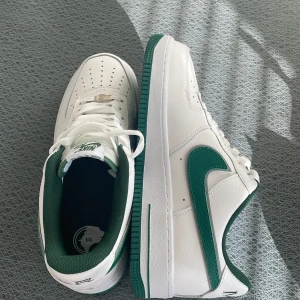 Nike Air Force i vitt och grönt - Snygga Nike Air Force sneakers i vitt med gröna detaljer. Skorna har en klassisk design med snörning och en grön sula. Perfekta för en stilren look med en touch av färg. (Egen design) va lite förstora och gick inte att lämna tillbaks eftersom det är egen design.