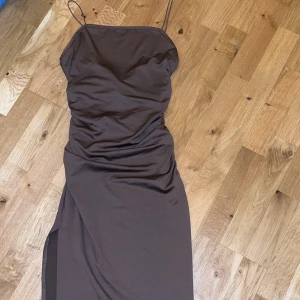 Brun klänning med smala axelband - Elegant brun klänning med smala axelband. Klänningen har en stilren design med en subtil drapering framtill och en slits på sidan. Även halvöppen rygg men korsning. Knälång för mig som är 175cm i storlek S