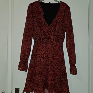 Röd och svart klänning från H&M Divided - Snygg röd och svart klänning från H&M Divided med ett djärvt leopardmönster. Klänningen har långa ärmar och volangdetaljer vid halsen och fållen. Perfekt för en trendig look!