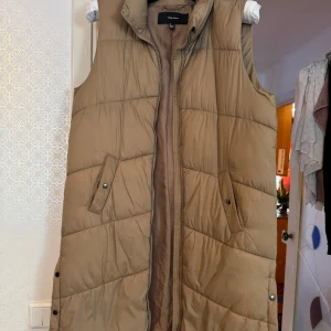 Beige dunväst från Vero Moda - Säljer en beige dunväst från Vero Moda. Västen har en dragkedja framtill och praktiska fickor med knappar. Perfekt för kyligare dagar och ger en stilren look.