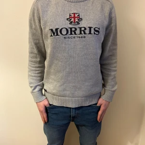 Grå tröja från Morris - Säljer en stilren grå tröja från Morris med ett broderat emblem och texten 'Morris Since 1968' på framsidan. Tröjan har långa ärmar och ribbade muddar vid ärmslut och nederkant. Perfekt för en klassisk look. Har storlek L men passar M