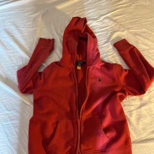 Röd huvtröja från Ralph Lauren - Säljer en snygg röd huvtröja från Ralph Lauren med dragkedja och broderad logga på bröstet. Tröjan har långa ärmar och en praktisk huva. Perfekt för en avslappnad stil.