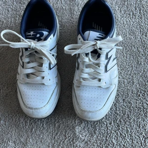 Vita sneakers från New Balance - Snygga vita sneakers från New Balance med marinblå detaljer och klassisk snörning. Perfekta för en avslappnad stil. Den ikoniska 'N' loggan pryder sidan av skorna. Perfekta till vår och sommar.