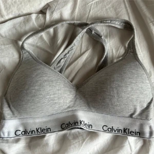 Sport BH Calvin Klein - Säljer en grå sport-bh från Calvin Klein med korsade axelband och elastiskt band med logotyp. Använt fåtal gånger💕