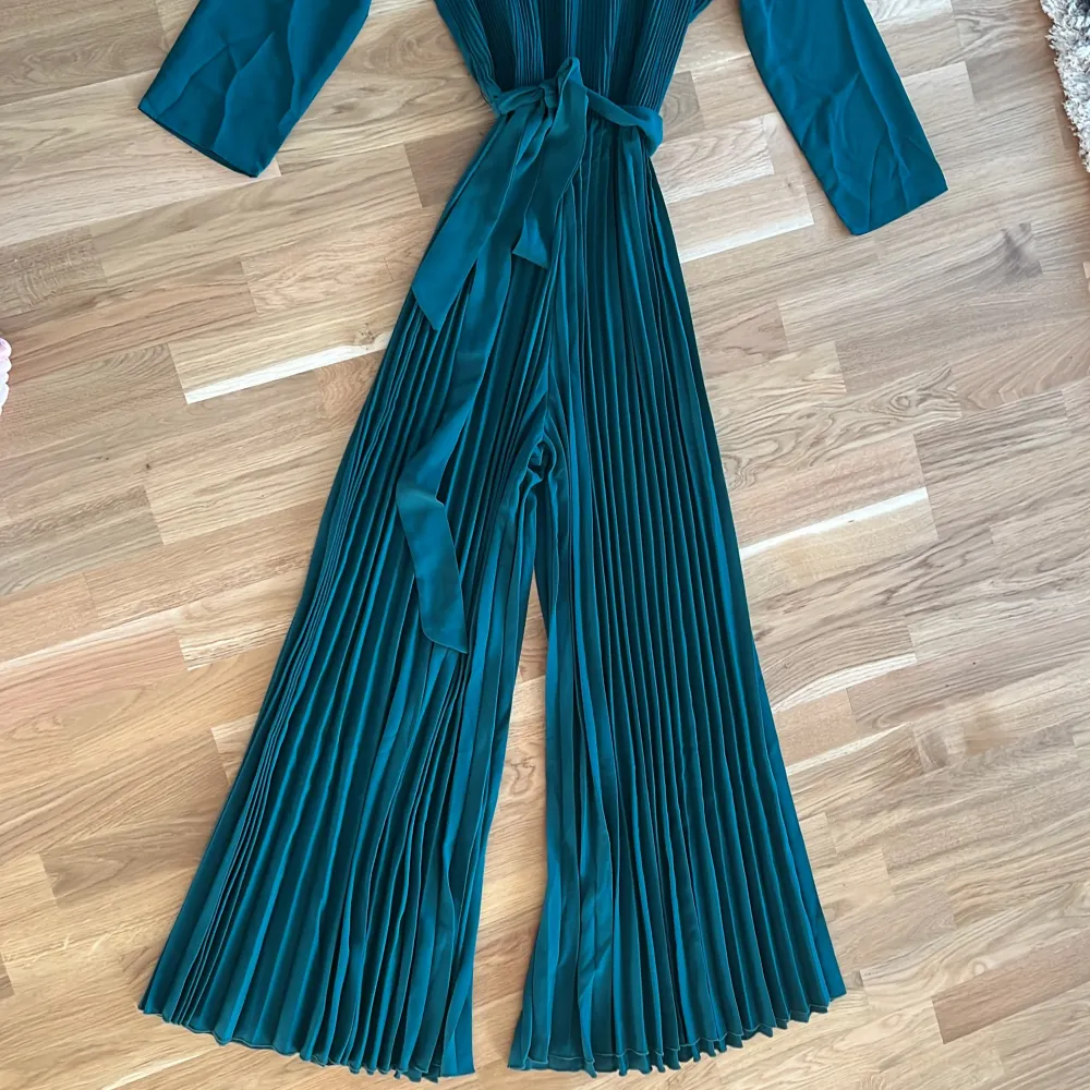 Snygg grön jumpsuit med plisserade ben och knytband i midjan. Den har trekvartsärmar och en hög krage som ger en elegant touch. Perfekt för en stilfull look.. Mekot.