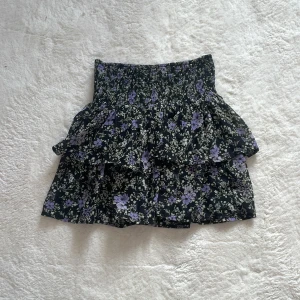 Blommig kortkjol med volang - Charmig kortkjol med blommigt mönster i lila och grönt på en svart bakgrund. Kjolen har en volangdetalj som ger extra flärd och en elastisk midja för bekväm passform. Perfekt för en somrig look!