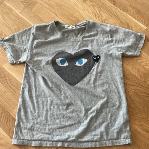 Grå t-shirt från Comme des Garçons - Säljer en grå t-shirt från Comme des Garçons med ett stort svart hjärta och blå ögon på framsidan. Den är kortärmad och perfekt för en avslappnad stil. Passar bra till både jeans och shorts!